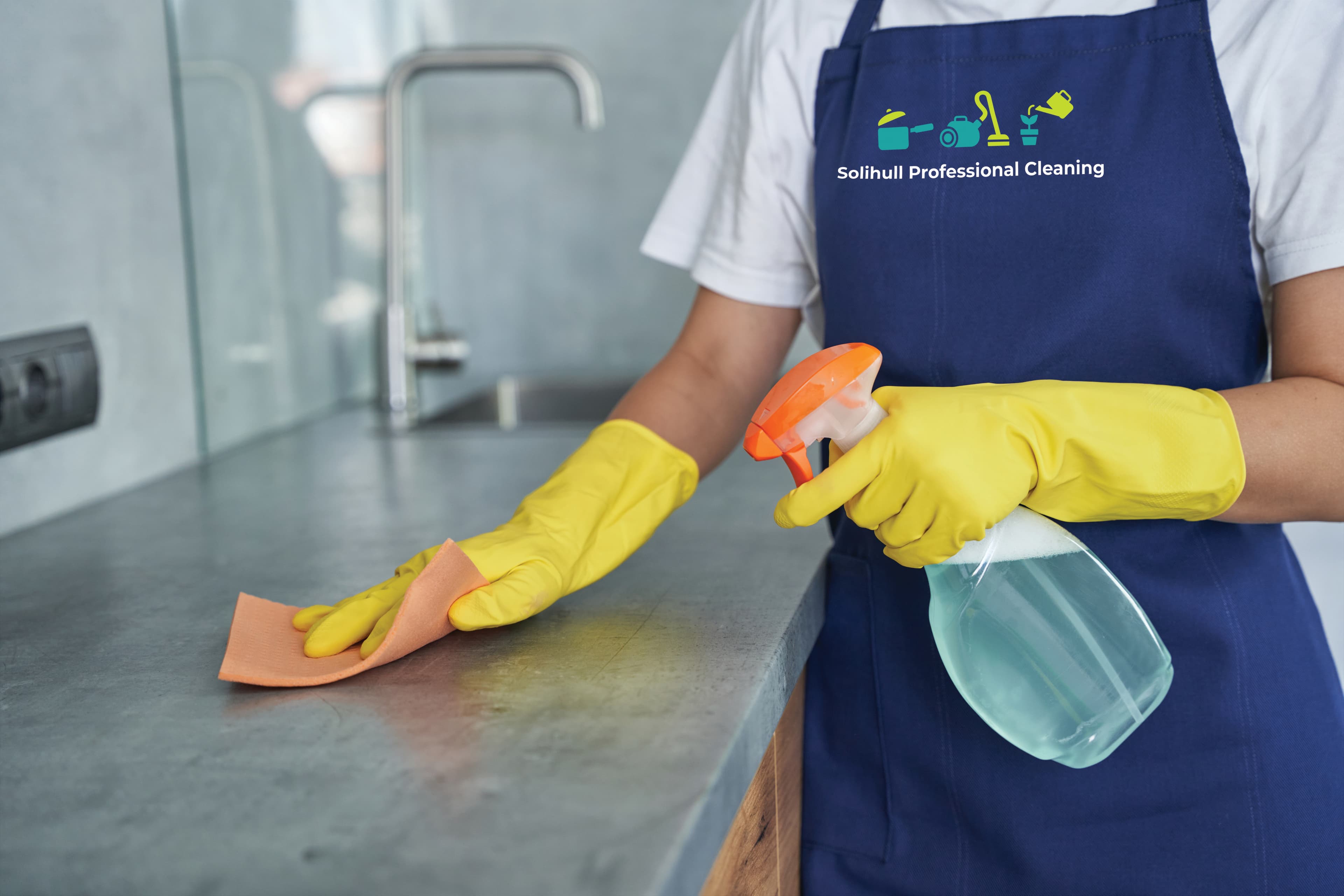 Cleaner in yellow gloves spraying and wiping a kitchen worktop